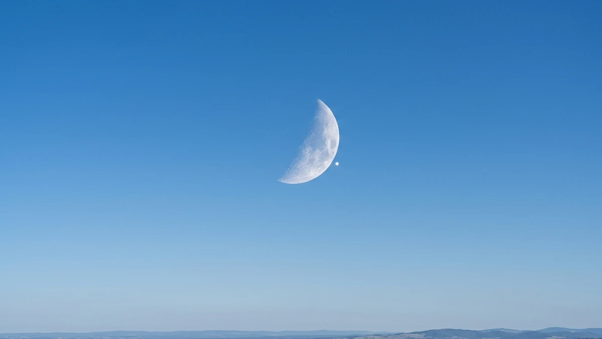 The crescent Moon against a blue daytime sky with the bright point of Venus about to disappear behind its dark edge