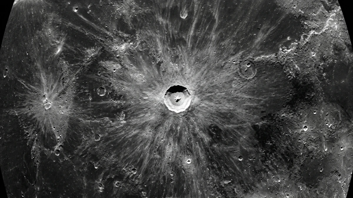 An overhead view of a small fresh bright crater with rays of ejecta on the grey lunar surface