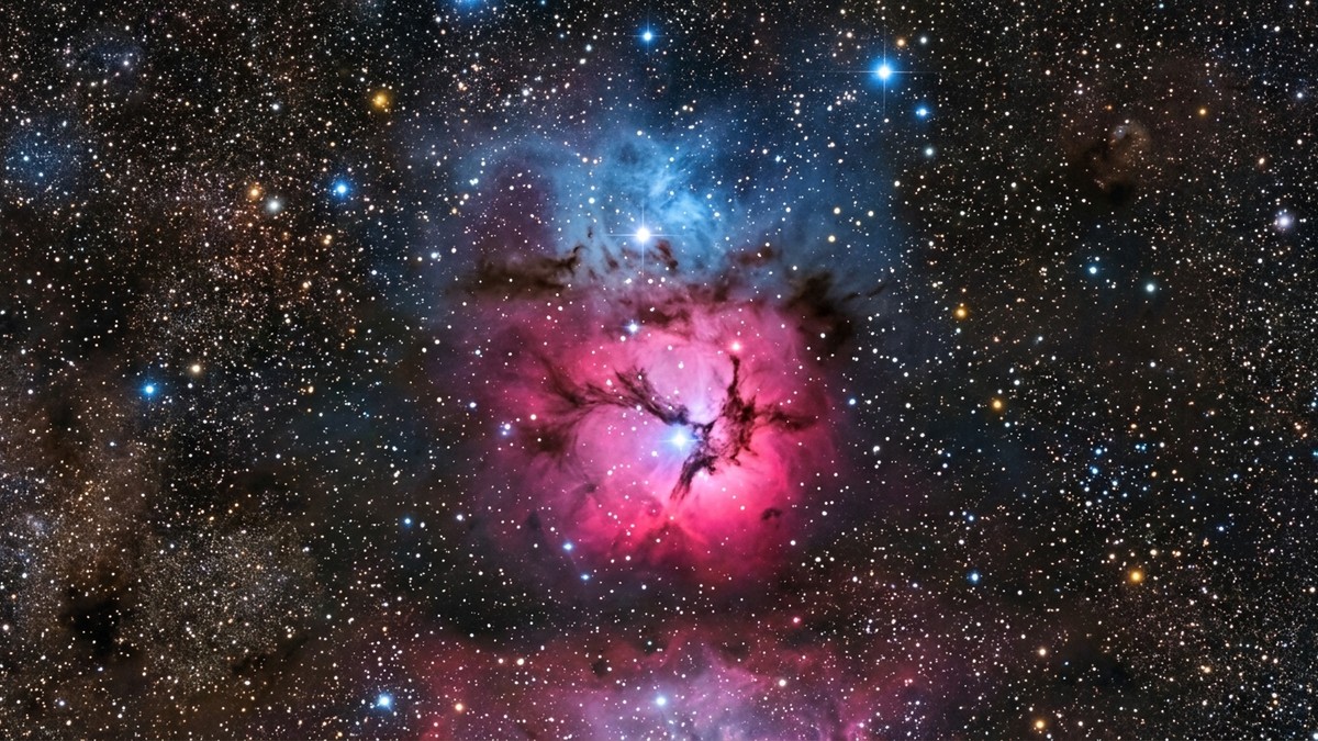 Wide view of the Trifid Nebula showing its three-lobed structure with pink emission, blue reflection, and dark dust lanes