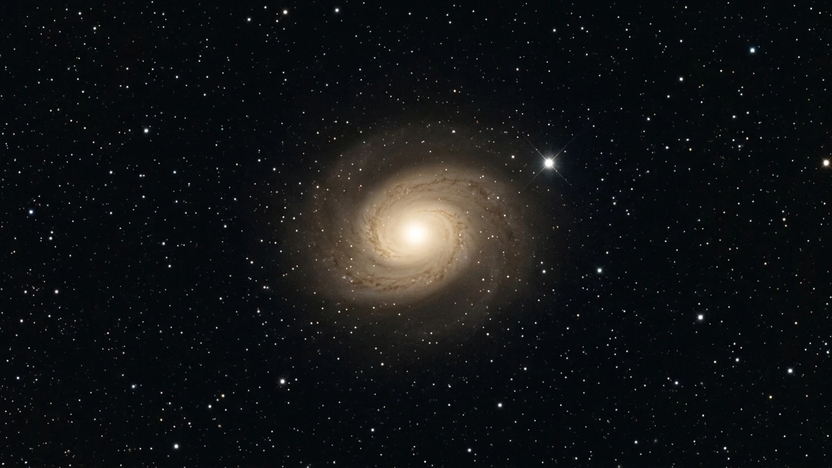 Visible-light image of spiral galaxy M77 (NGC 1068) showing its bright nucleus and faint spiral arms in pale gold against a deep black starfield, as it appears through ground-based telescopes