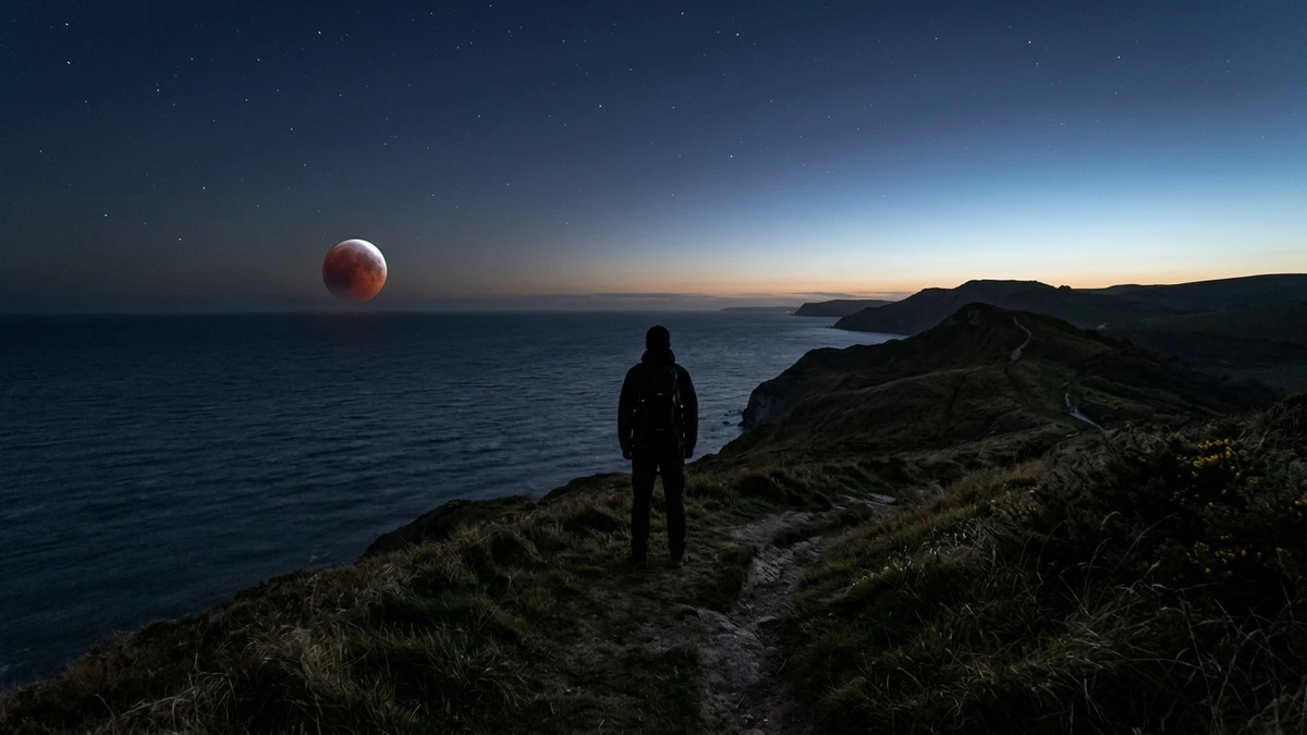 A lone observer on a west-facing UK coastal headland watching the eclipsed red Moon low over the sea before dawn
