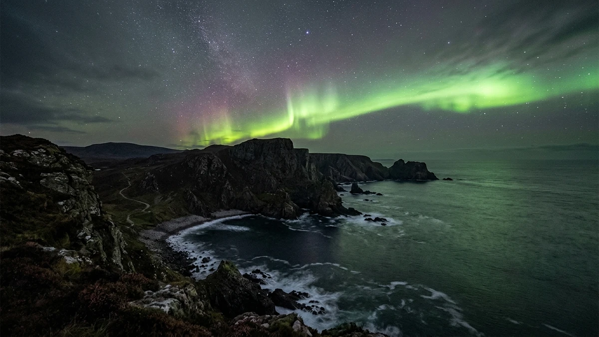 Brilliant green northern lights over a rugged Scottish coastline with dark cliffs and ocean below