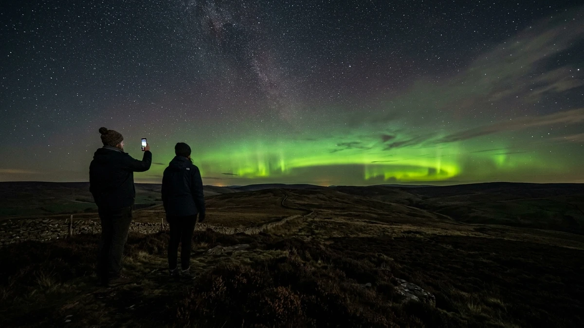 Two people standing on a dark hillside watching green northern lights on the horizon, one holding up a phone to photograph the display