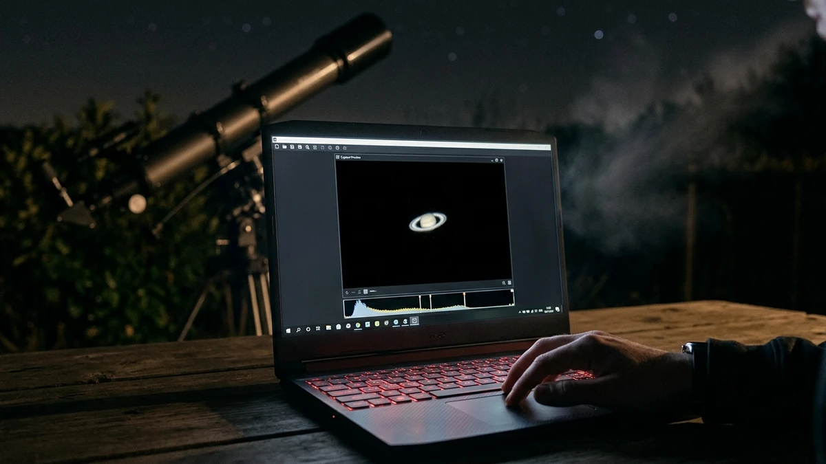 A laptop screen at night showing a live capture preview of Saturn with a histogram beneath it, telescope out of focus behind