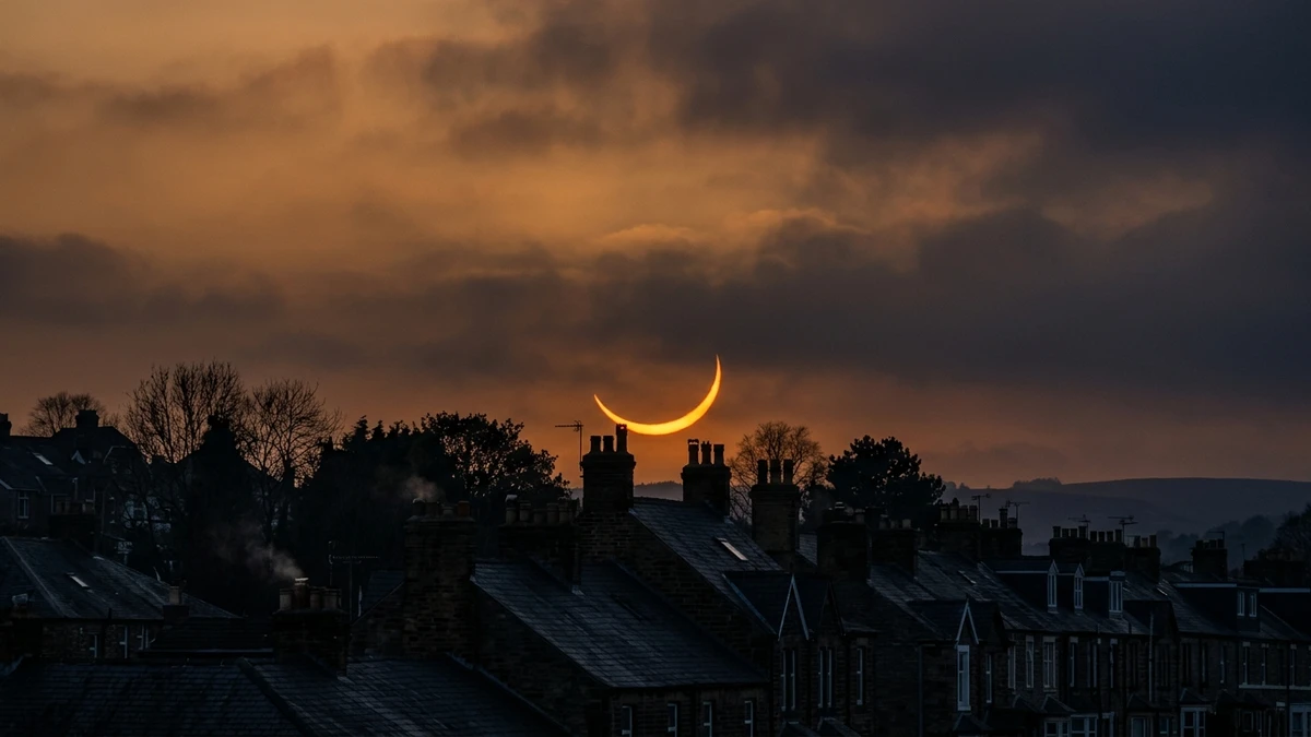 A thin crescent sun during a deep partial solar eclipse glowing orange low above rooftops and trees