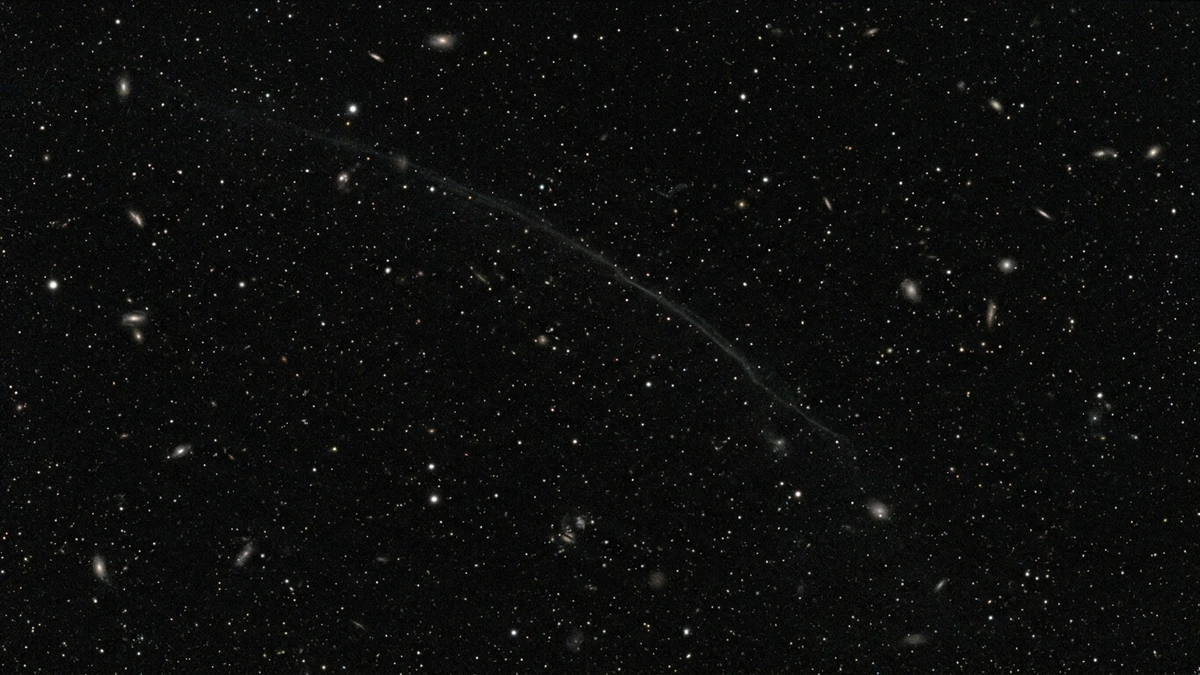 A faint stellar stream picked out as a curved ribbon of stars against the dim glow of a distant galaxy in a deep telescope image