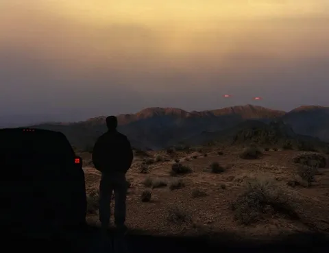 A digital rendering of a person standing next to a vehicle, looking at two red lights in the sky over a mountainous landscape.
