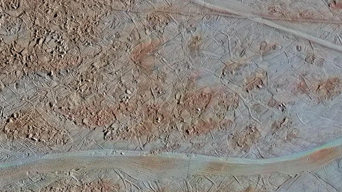 Enhanced-colour view of Europa's surface showing broken blocks of chaos terrain in pink-brown and a long, wide pale band running across the lower part of the frame