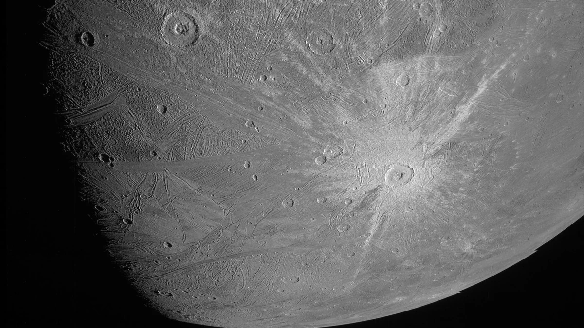 Ganymede in black and white from Juno, with a large crater and bright rays right of centre and grooved ground to the left