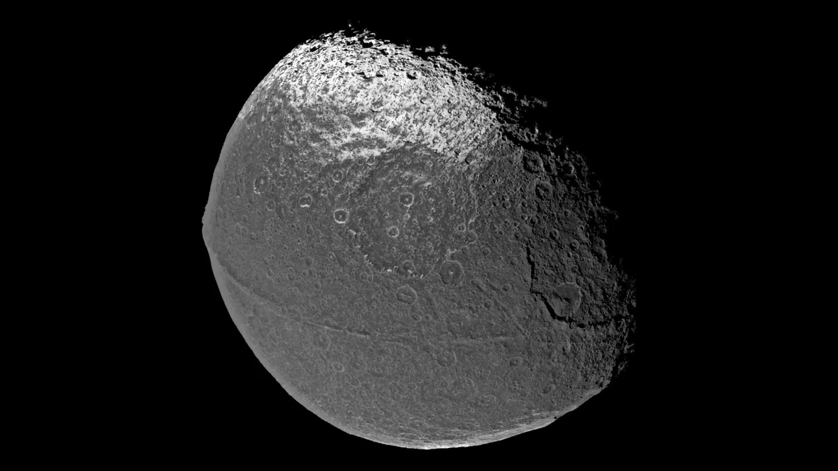 Iapetus photographed by Cassini, a dark heavily cratered disc with a thin ridge running across its equator from the left edge towards the shadow on the right