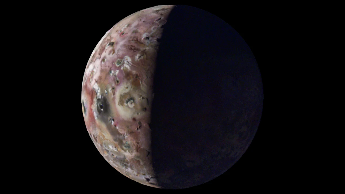 Io photographed by Juno's JunoCam, a sunlit crescent of pink, cream and brown ground dotted with dark volcanic pits and pale deposits on the left, with the night side fading into black on the right