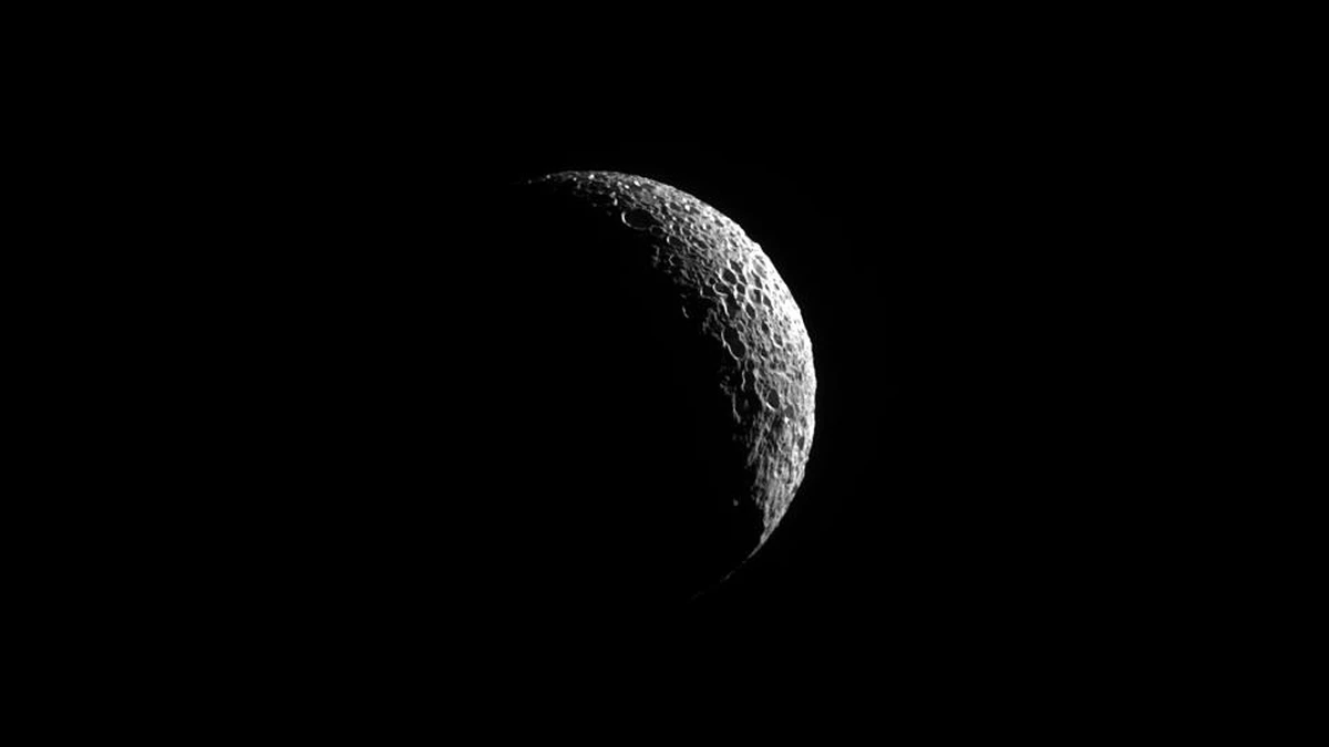 A thin bright crescent of Mimas in black and white against black space, with long shadows picking out rows of overlapping craters along the lit edge